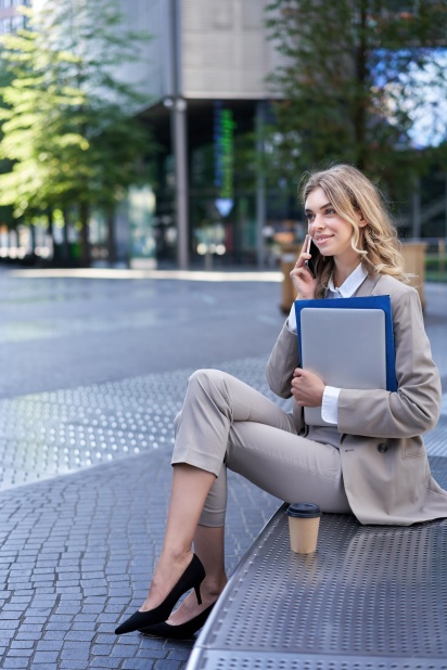 Vertical shot of young woman sitting near office building, holding laptop and folders with work documents, calling someone on mobile phone.